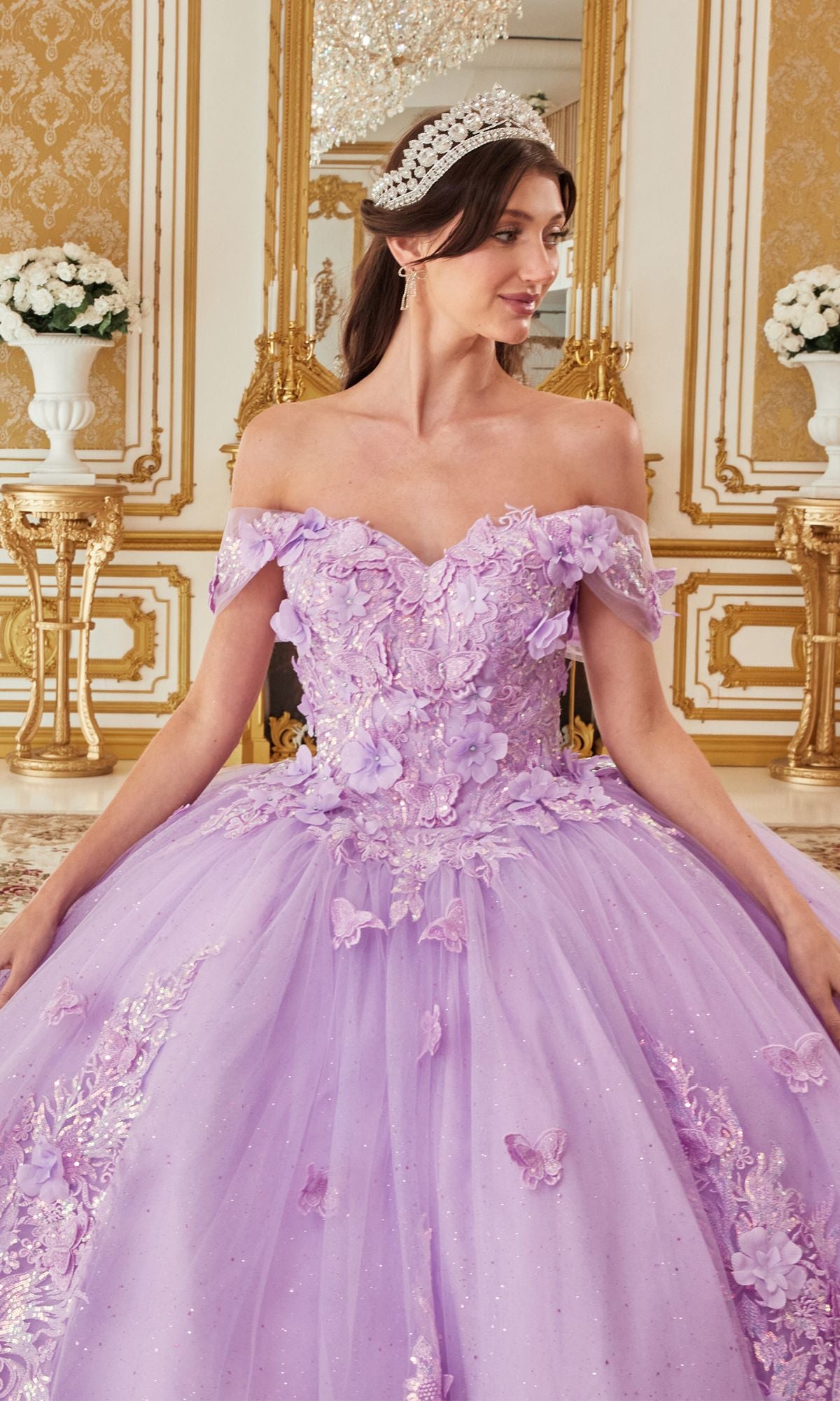 A woman in the Quinceanera Dress 15713 by Ladivine, featuring off-the-shoulder styling with butterfly appliqués and floral accents, stands in a lavish gold-decorated room, her tiara sparkling above the elegant lavender ball gown.